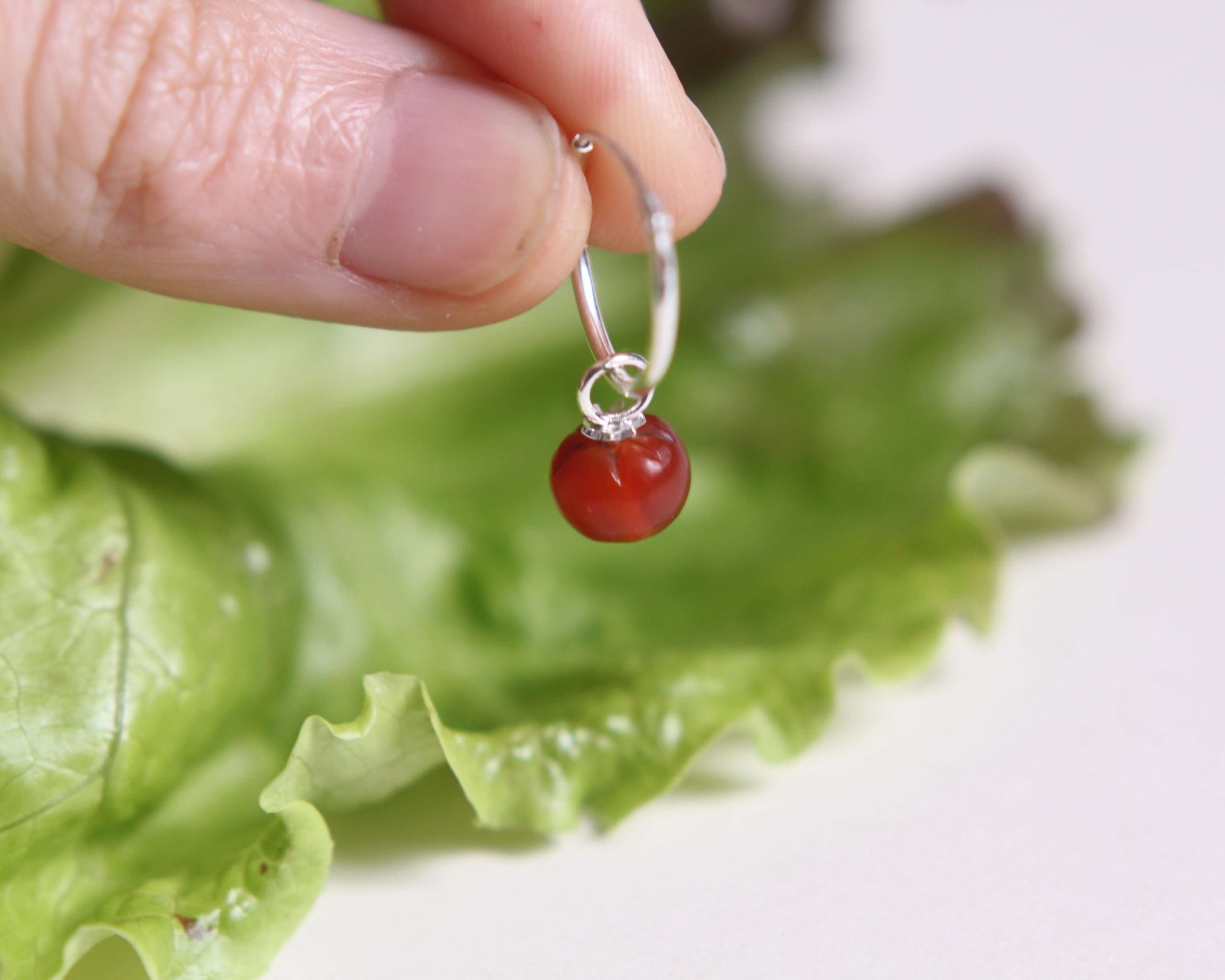 Pendentif tomate sur boucle d'oreille tenue par une main en argent posé sur une feuille de salade sur un fond blanc