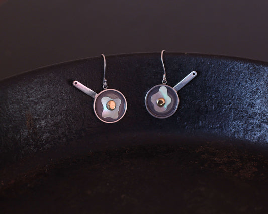 paire de boucles d'oreilles symbolisant des oeufs au plat, accrochée au rebord d'une vraie poêle