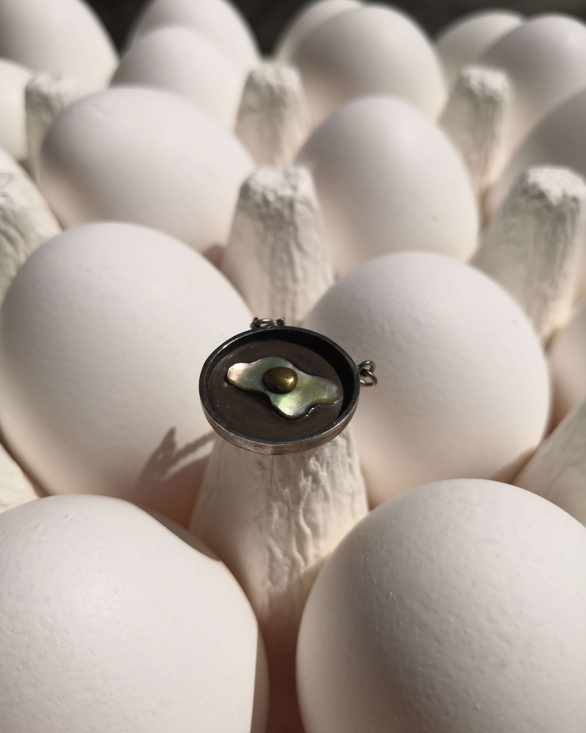 un pendentif en argent en forme d'oeuf au plat en nacre et en laiton est posé dans une barquette d'oeufs