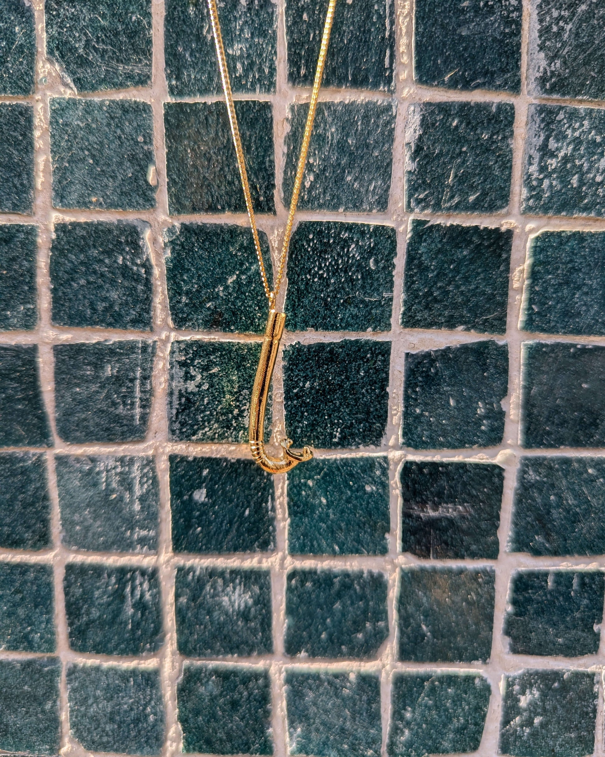 collier avec un pendentif en laiton plaqué or en forme de tuba devant un mur à carreaux